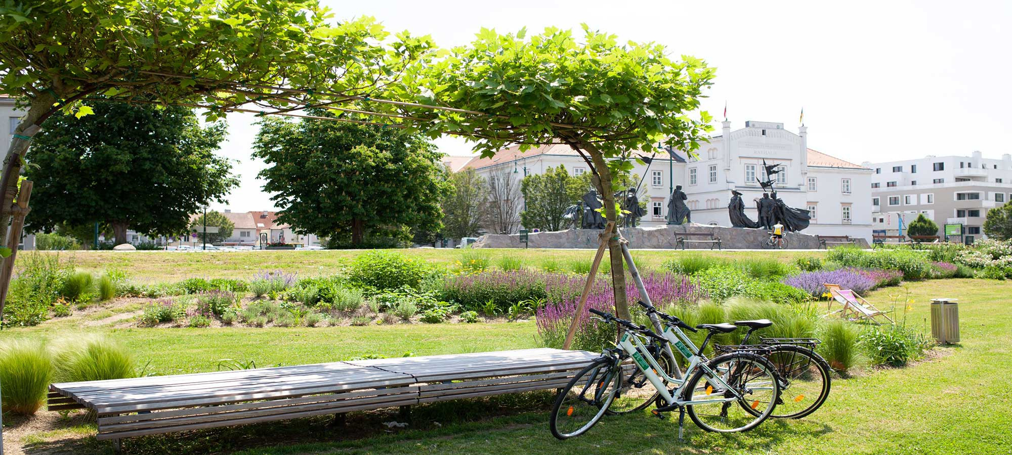 Fahrräder unter Laubengang in gepflegtem Park mit Blick auf historisches Gebäude und Statue im Hintergrund