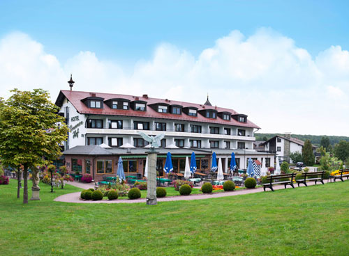Hotel mit Außensitzbereich, umgeben von Bäumen und Grün unter blauem Himmel.